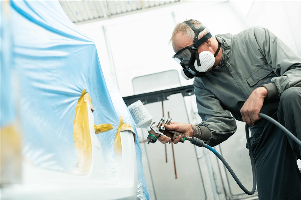 Schutzmaske bei Arbeit mit Lackierung Ein Mann trägt eine Atemschutzmaske beim Sprühen von Farbe in einem Lackierraum.