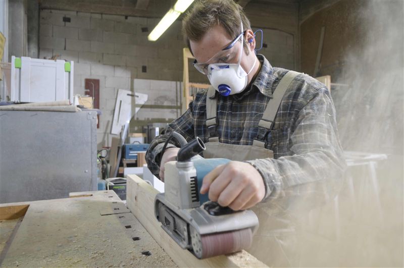 Sicherheitstechnische Schleifarbeit Mann schleift Holz mit einem elektrischen Bandschleifer in einer Werkstatt.