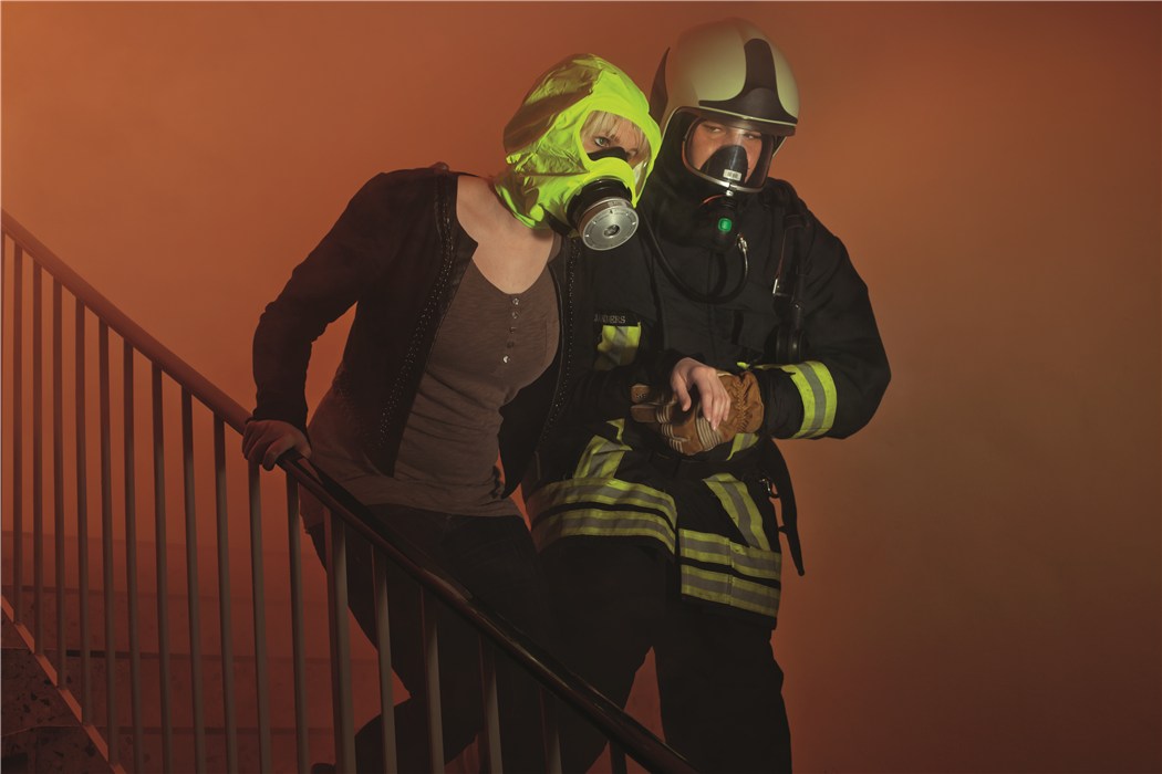 Evakuierung mit Feuerwehratemschutz Feuerwehrmann mit Atemschutzmaske begleitet Person mit Fluchthaube die Treppe hinunter in rauchiger Umgebung.