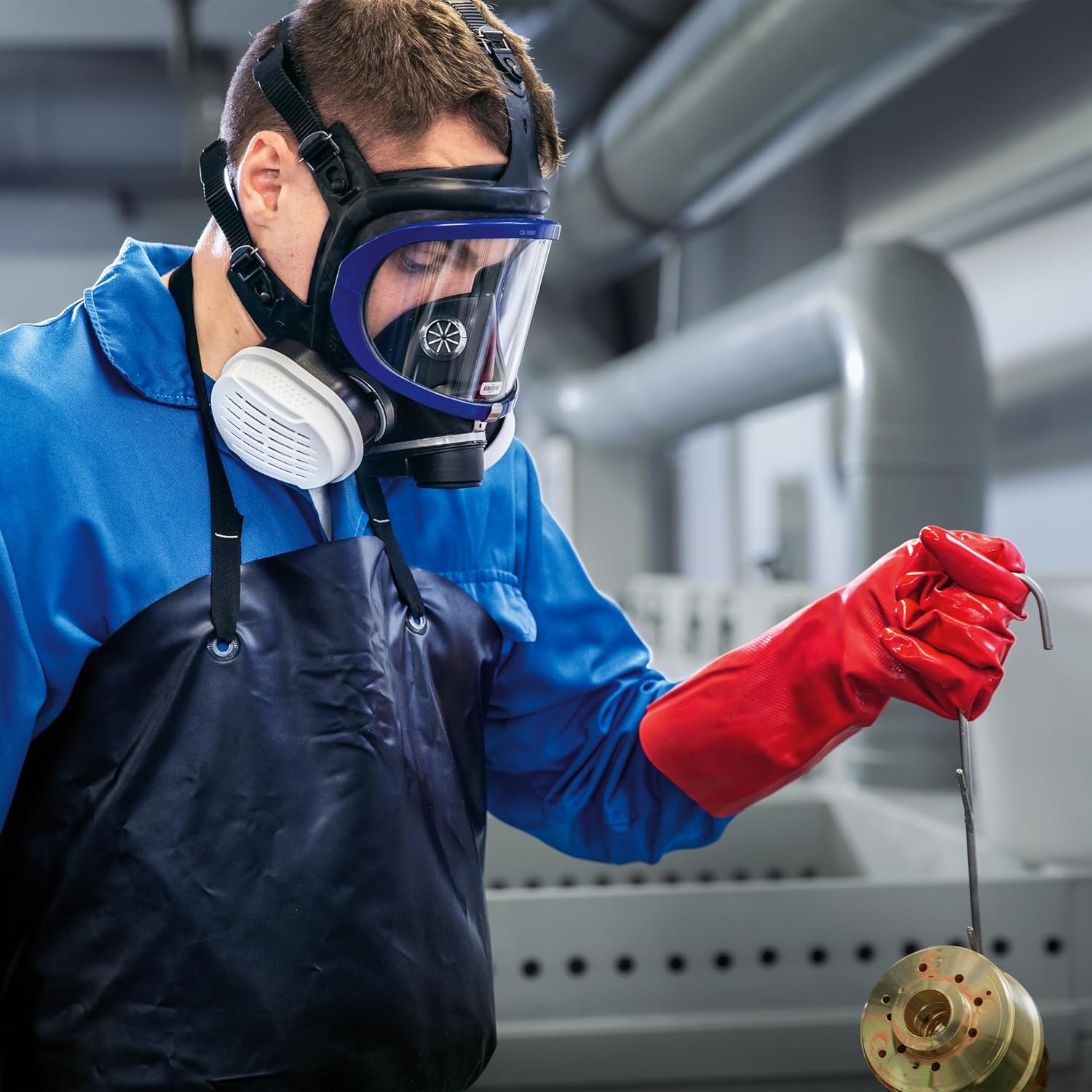 Vollmaske Dräger X-plore 5500 in Aktion Person trägt Dräger X-plore 5500 Vollmaske, rote Handschuhe und blauen Schutzanzug in einem industriellen Umfeld.