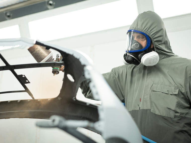 Schutzmaske für sichere Lackierungsarbeiten Person in Schutzanzug und Maske beim Lackieren eines Fahrzeugteils.