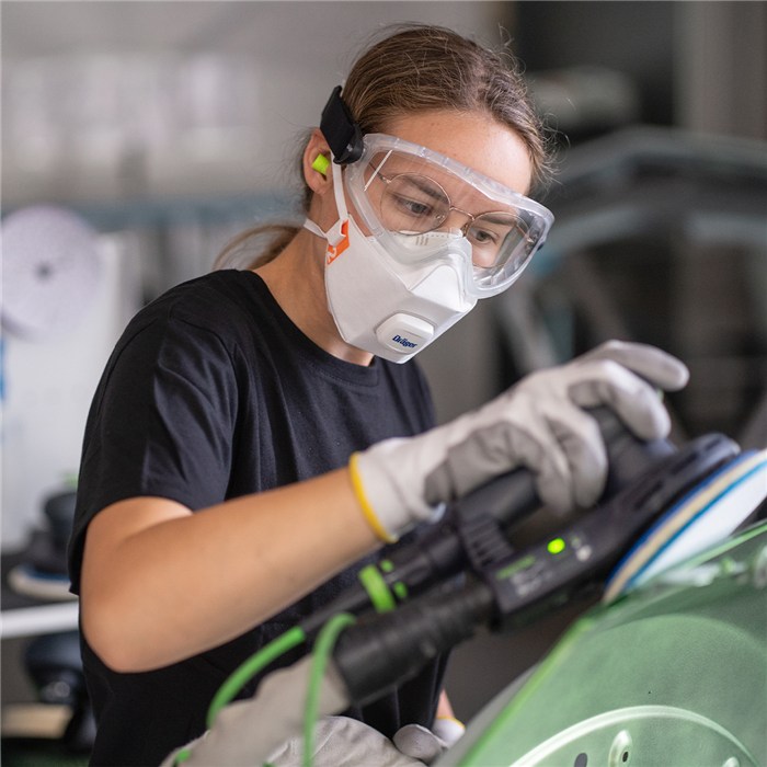 Schutzausrüstung bei der Fahrzeugaufbereitung Person trägt Gehörschutz und Handschuhe, arbeitet an grünem Fahrzeug mit Schleifgerät.